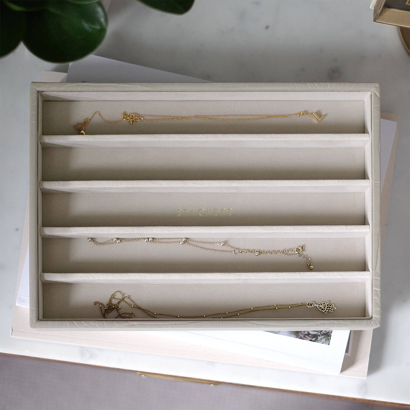 Putty croc necklace tray with gold necklaces inside the compartments, sitting on a stack of books on a table.