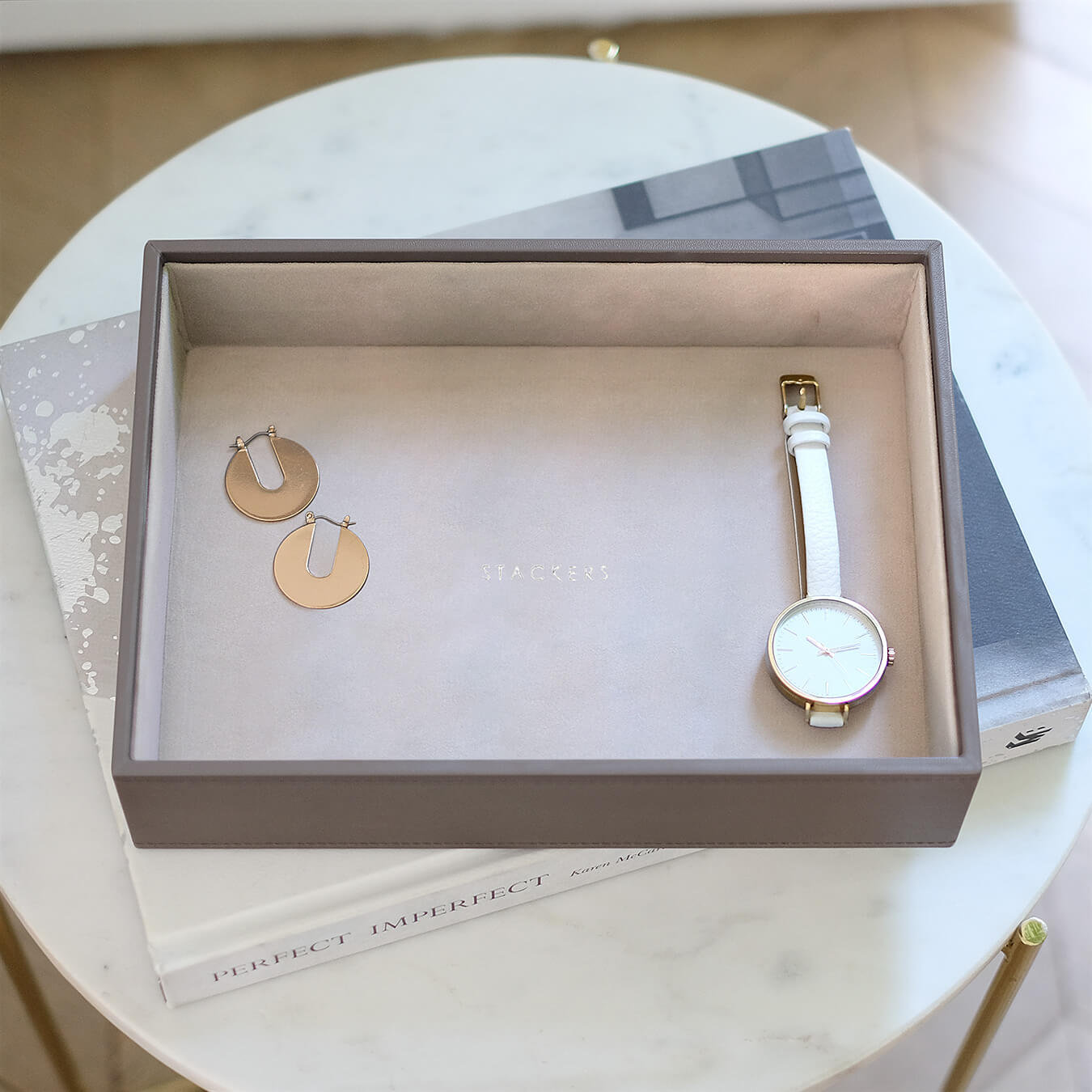 Gold hoop earrings and white watch in a mink jewelry box stacked on a book on a side table